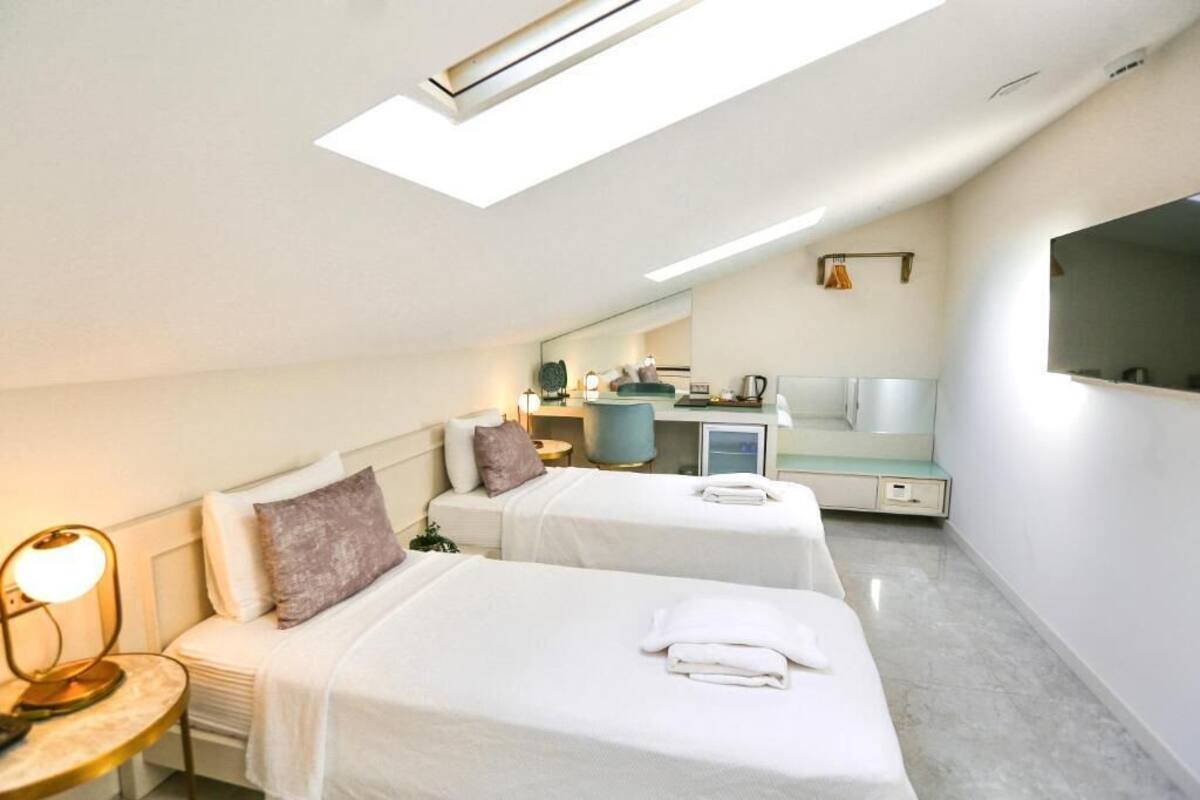 A hotel room with two beds, a desk, a chair, a lamp, a mirror, and a skylight.