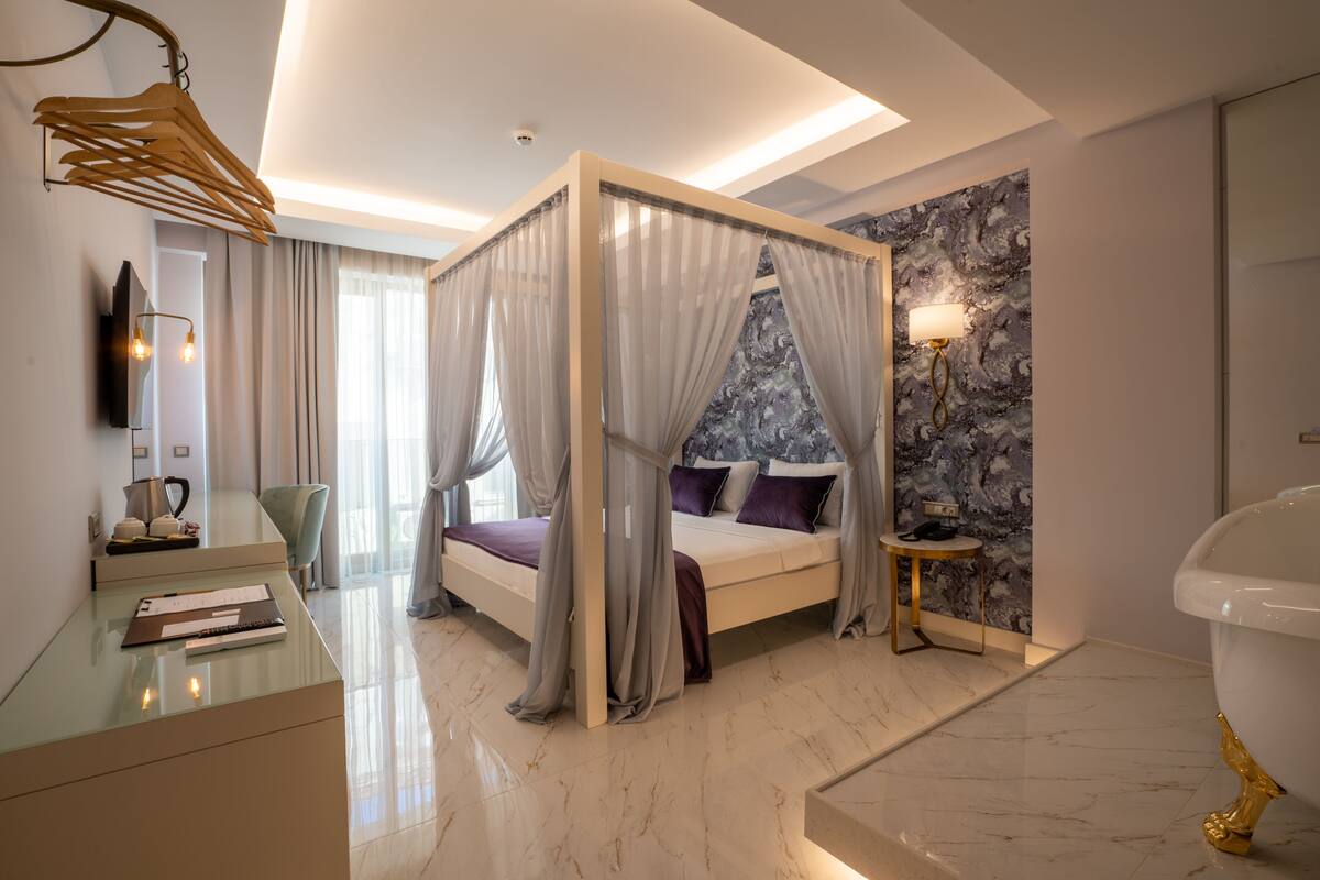 A modern bedroom with a four-poster bed, a bedside table, a chair, a vanity with a mirror, and a bathroom with a bathtub.