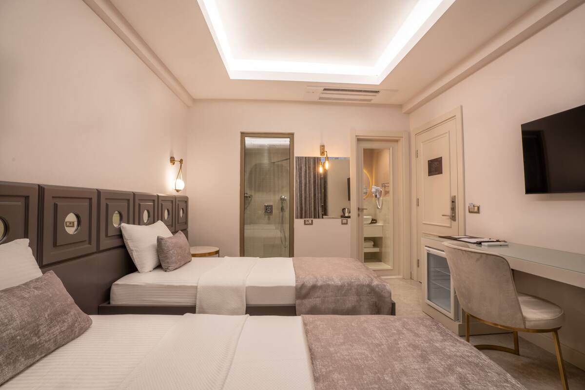 A hotel room with two beds, a desk, a chair, a TV, and a bathroom with a glass door.