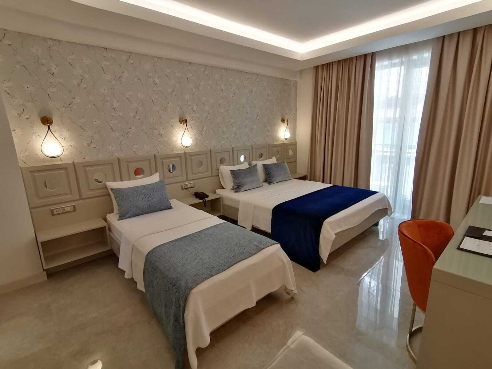 A hotel room with two beds, a desk, and a chair. There are wall sconces, a window with curtains, and a marble-patterned wall.