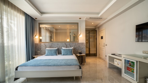 A modern hotel room with a large bed, a small bedside table, a built-in refrigerator, and a large mirror above the bed. - Muhtar Hotel (Kas)