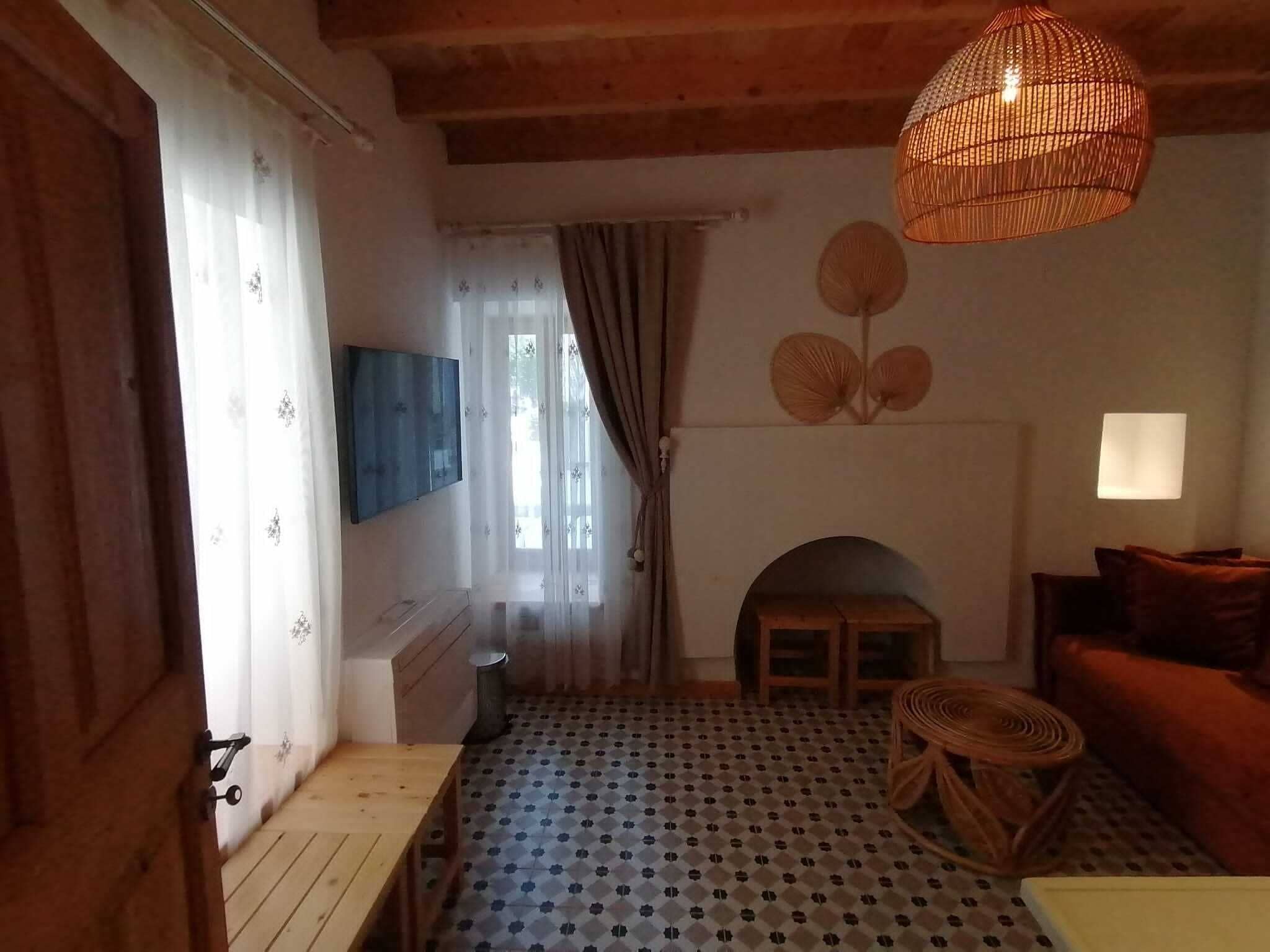 A cozy room with a wooden ceiling, a patterned floor, a sofa, a table, a hanging light fixture, and a fireplace.