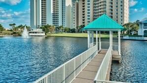 A wooden dock leading to a gazebo with a green roof, overlooking a calm body of water with a fountain in the background and high-rise buildings in the distance. - Home with Pool in the Gated Community of Caribe (Destin)