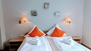 Habitación de hotel con dos camas, cada una con sábanas blancas y almohadas naranjas, y dos lámparas fijadas a la pared.