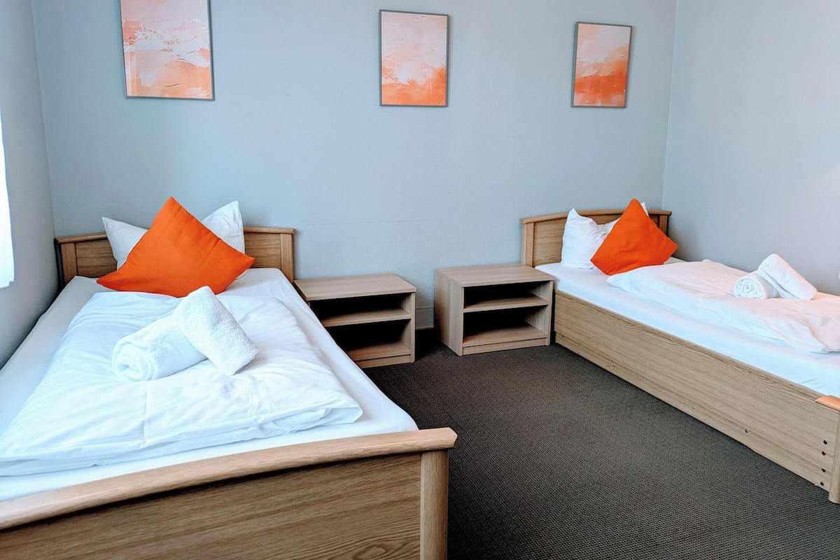A hotel room with two beds, each with white linens and orange pillows, and a small wooden nightstand between them.