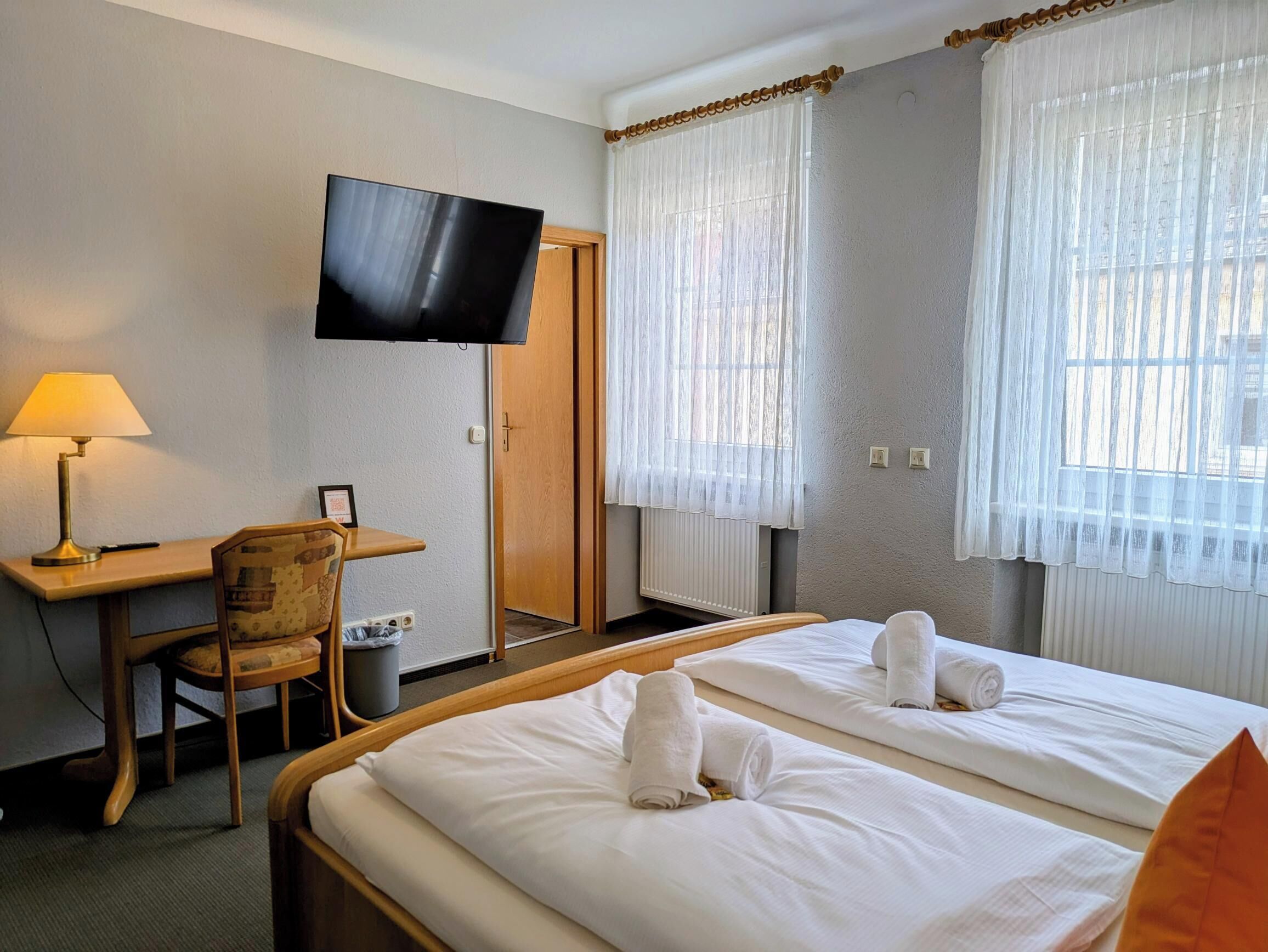 A hotel room with a bed, a desk, a chair, a television, and two windows with curtains.
