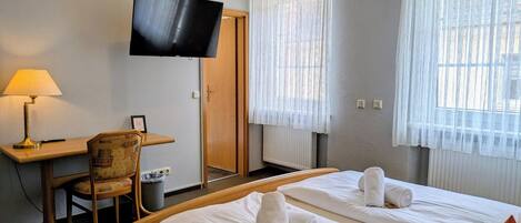 A hotel room with a bed, a desk, a chair, a television, and two windows with curtains.