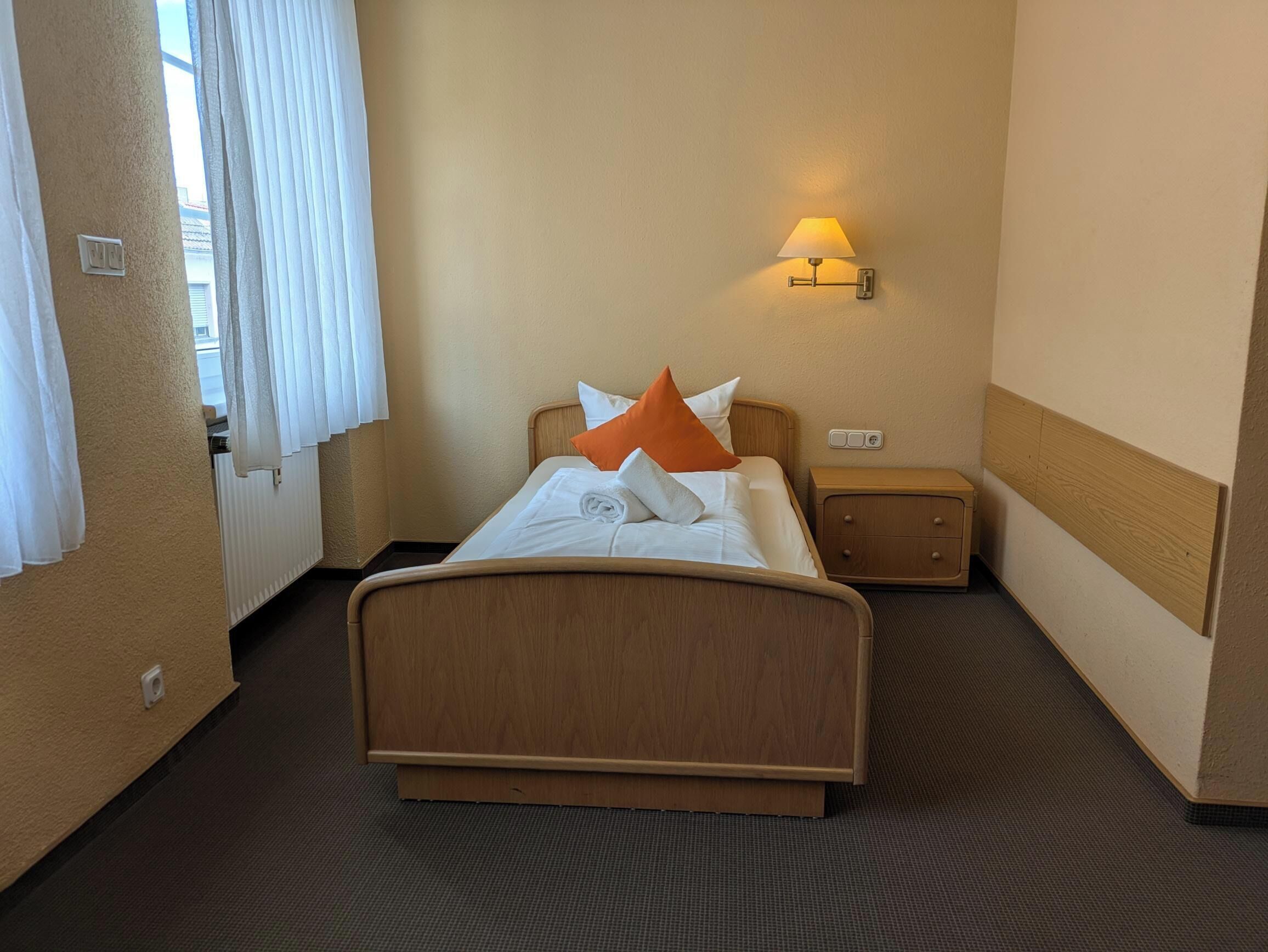 Ein Hotelzimmer mit einem hölzernen Bett, weißer Bettwäsche, einem orangen Kissen, einem Nachttisch und einer Wandlampe.