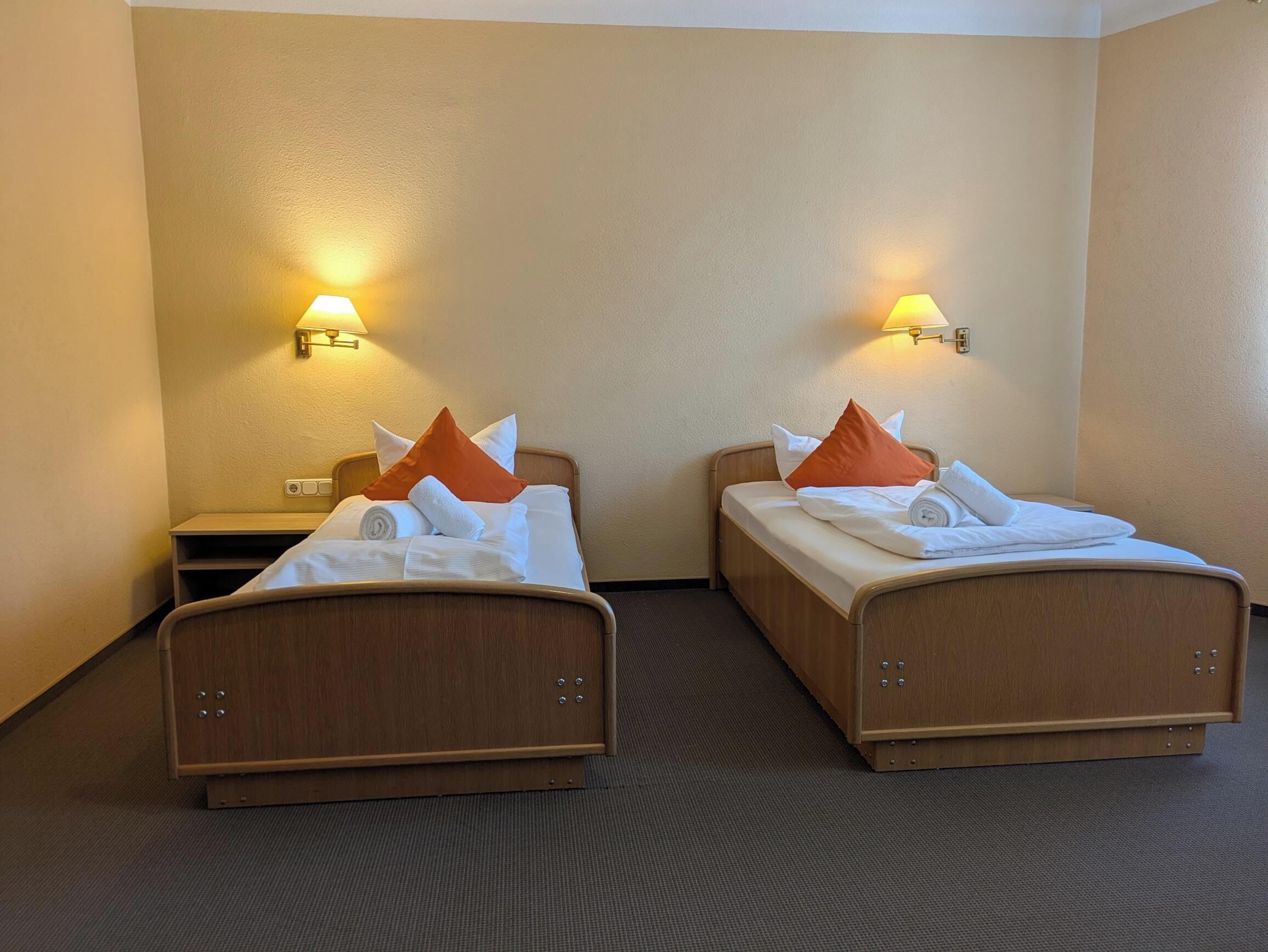 Ein Hotelzimmer mit zwei Einzelbetten, jeweils mit Holzrahmen und weißer Bettwäsche. Zwei Wandlampen sorgen für Beleuchtung.