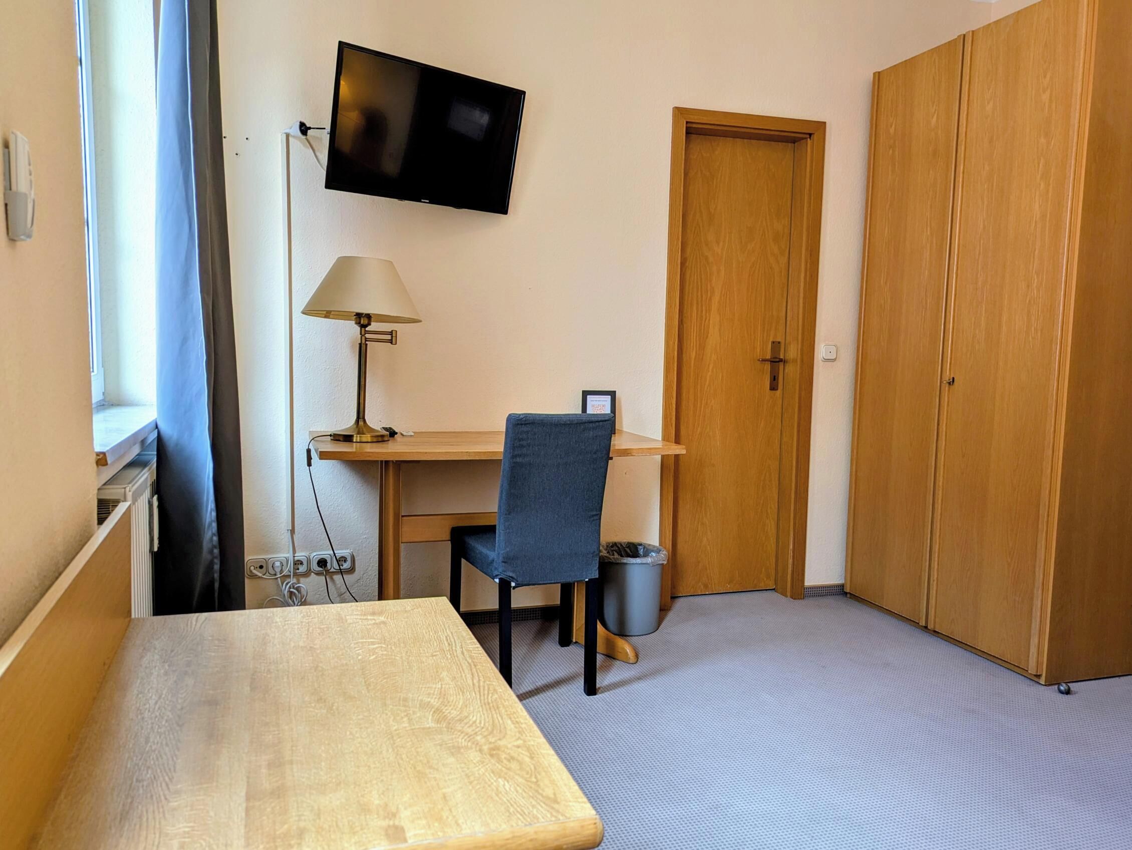 Ett litet rum med ett skrivbord i trä, en stol, en tv som är monterad på väggen, en lampa och en garderob.