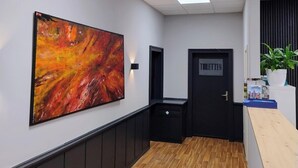 Un pasillo con suelo de madera, un cuadro abstracto grande en la pared, una puerta con la leyenda 'TOILETEN' y un mostrador de recepción con una planta.