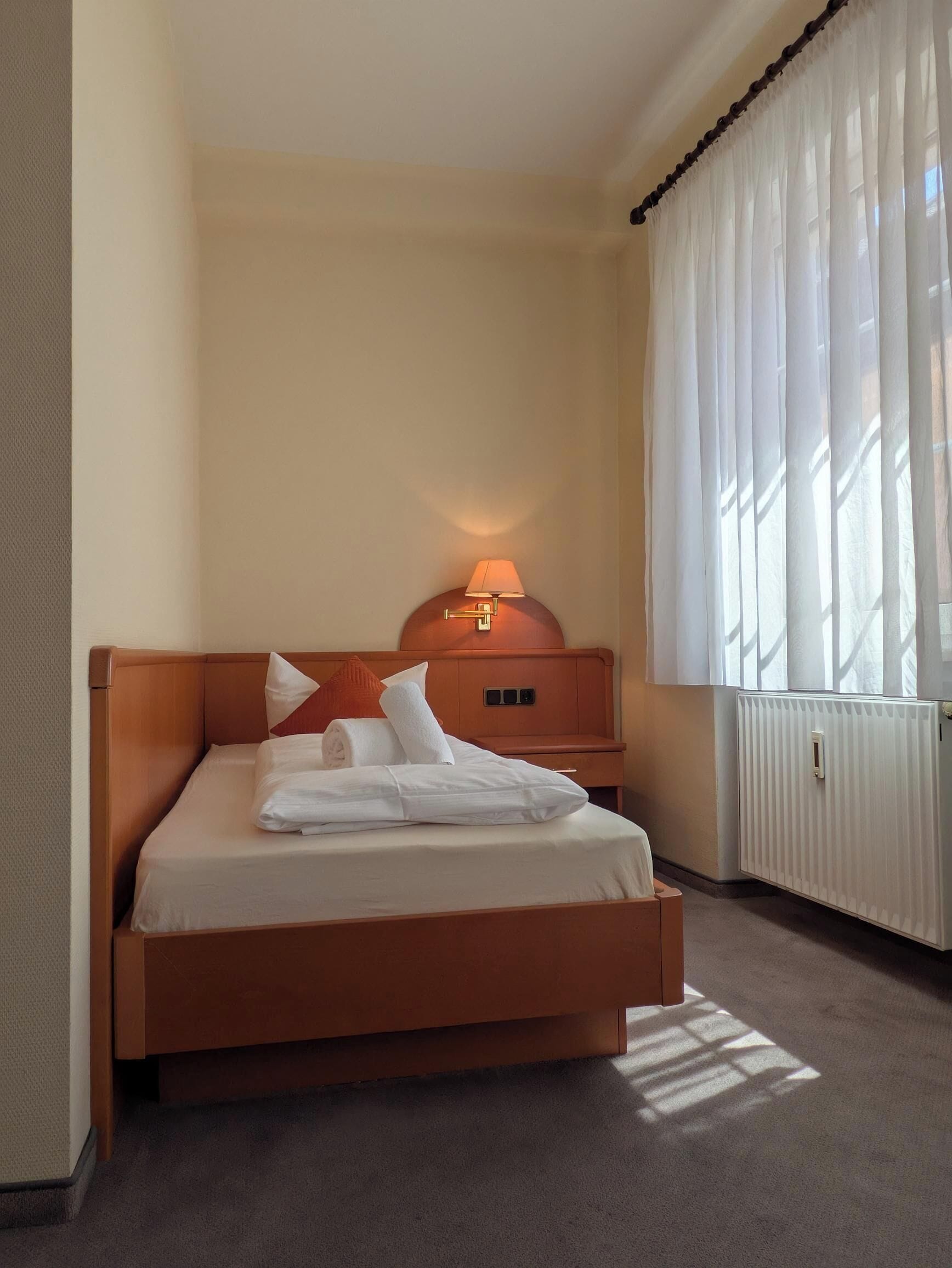 Ein Hotelzimmer mit einem Einzelbett, einem Nachttisch mit Lampe, einem Fenster mit durchsichtigen Vorhängen und einem Heizkörper.