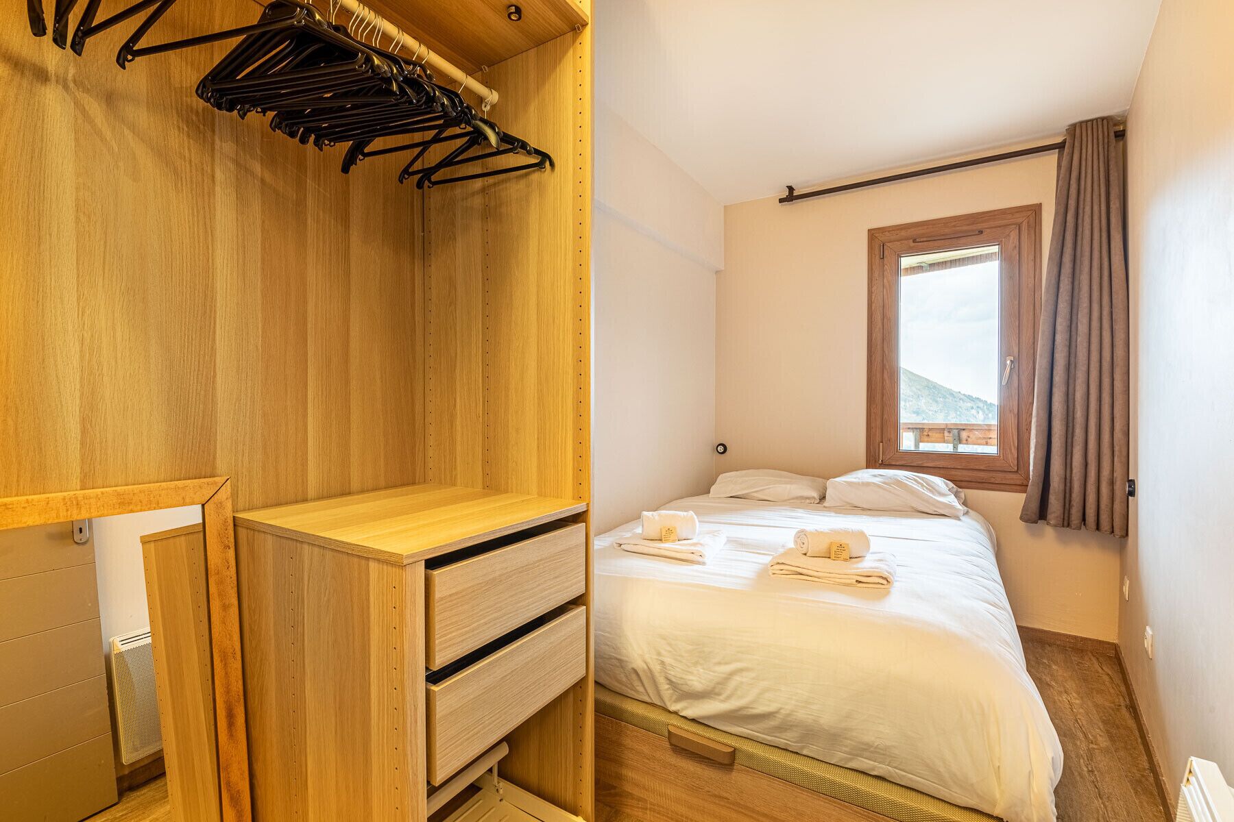 Ein gemütliches Schlafzimmer mit Holzschrank, einem Bett mit ordentlich gefalteten Handtüchern und einem Fenster mit Vorhängen.