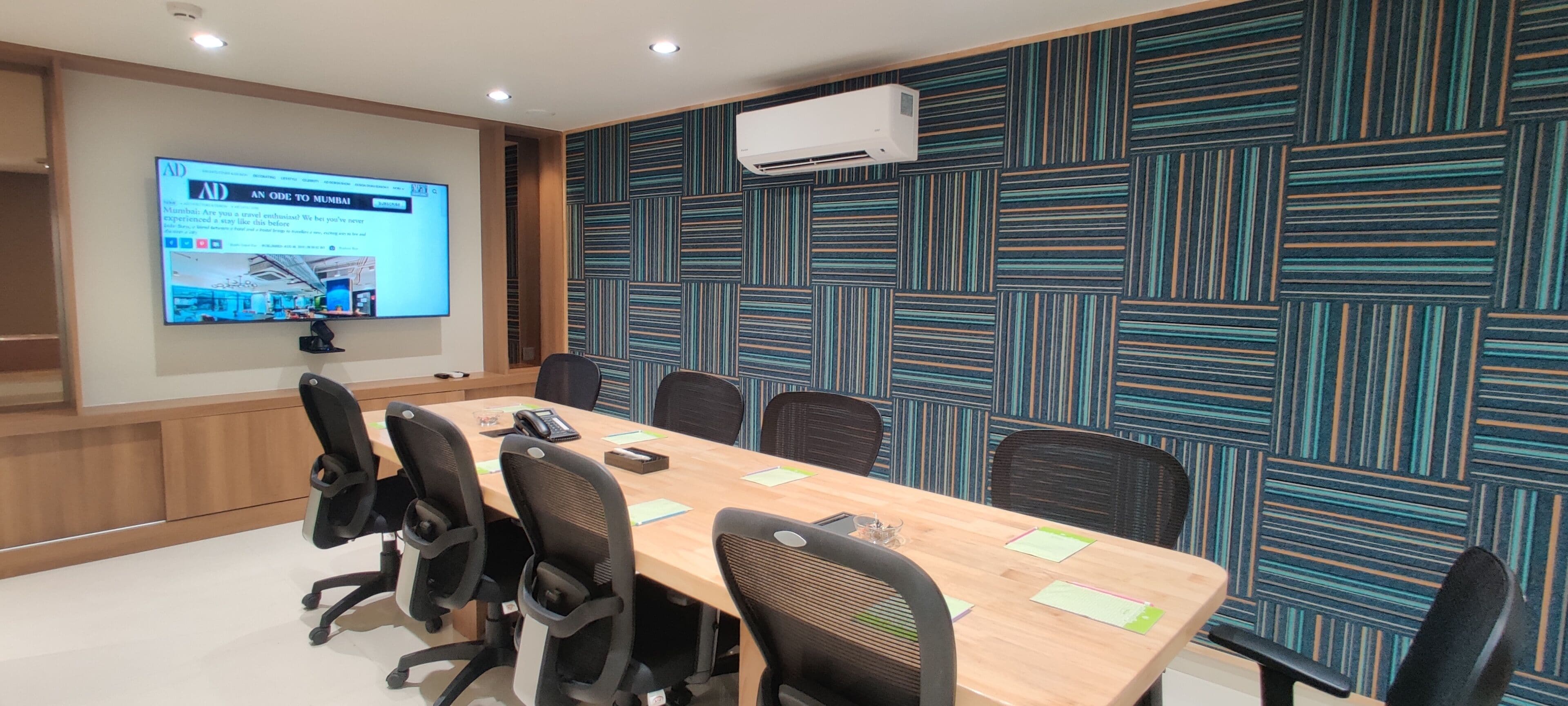 A conference room with a long table, six chairs, a projector screen displaying an advertisement, and a patterned wall.