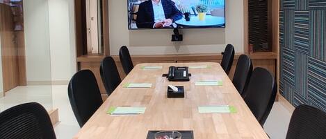 A conference room with a long table, black chairs, a mounted TV screen, and ceiling lights.
