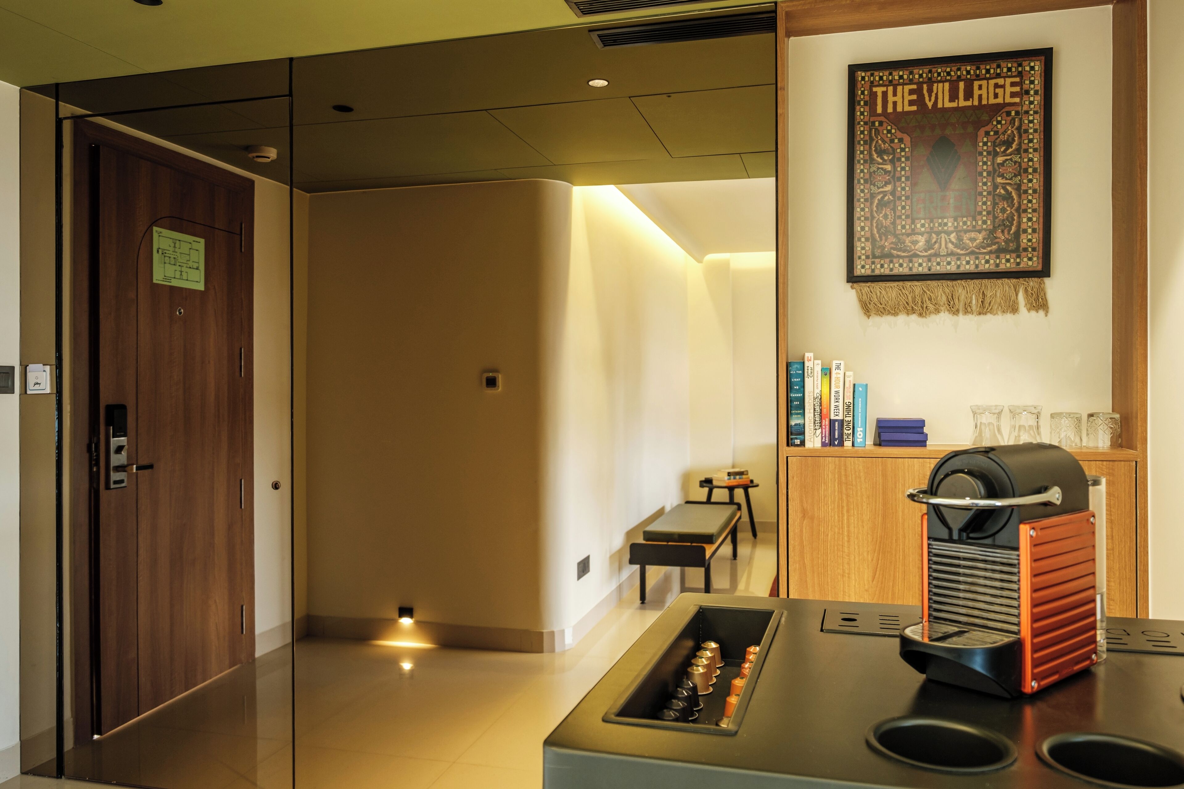 A hotel lobby with a wooden door, a bench, a coffee machine, and a wall-mounted sign reading "THE VILLAGE".