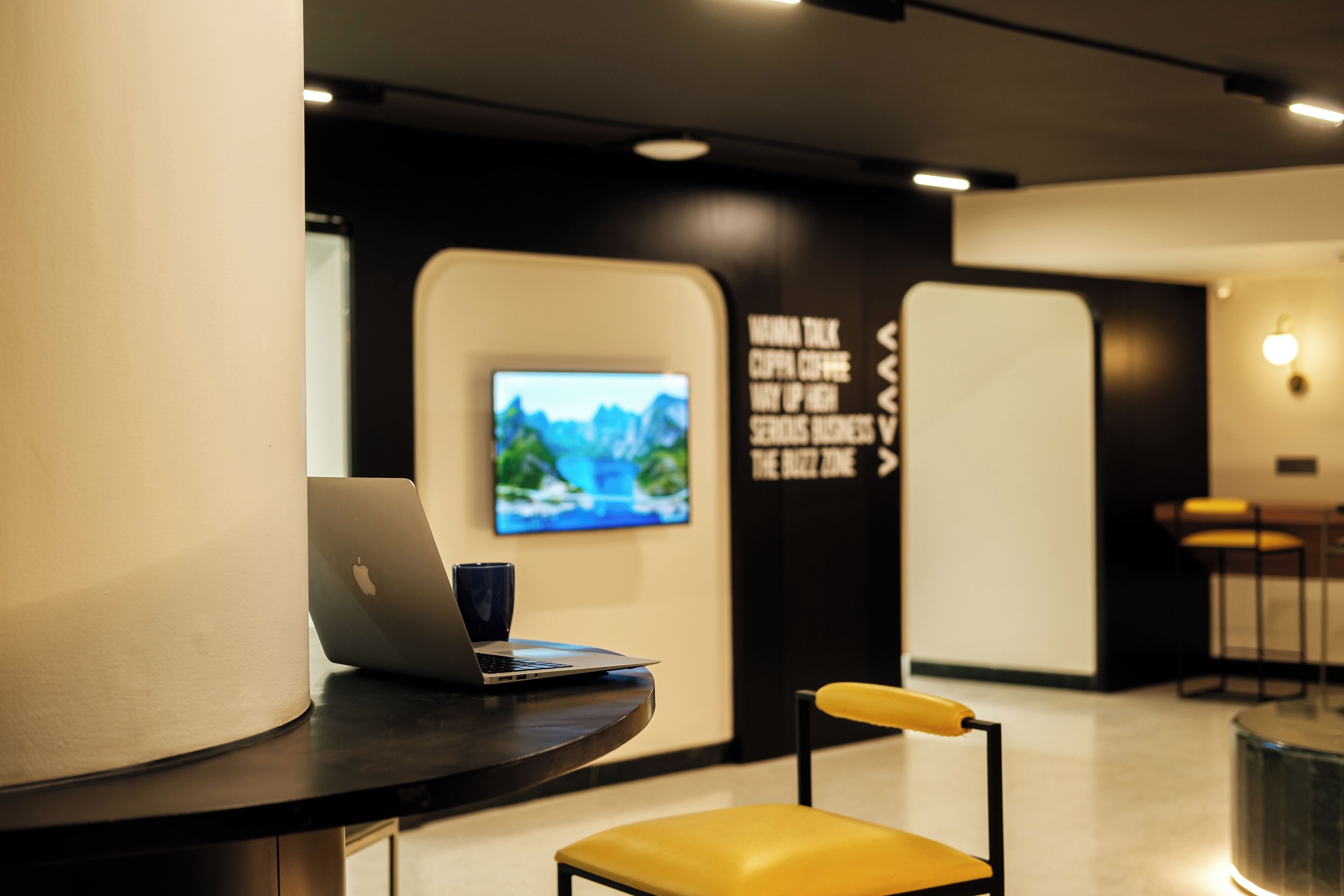 A modern workspace with a laptop on a curved table, a yellow chair, and a wall-mounted TV displaying a scenic image.