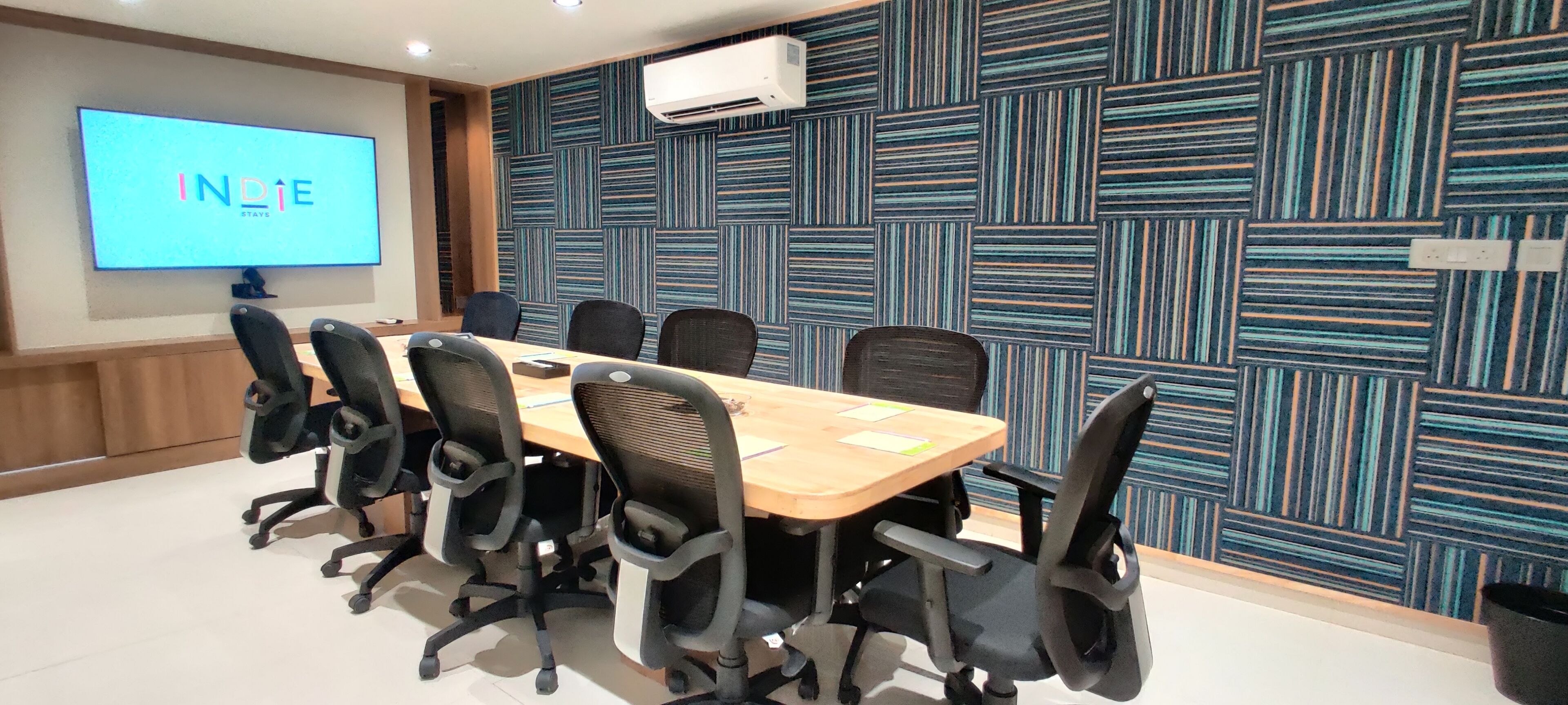 A conference room with a long table, black office chairs, a projector screen displaying "INDIE STAYS," and a patterned wall.