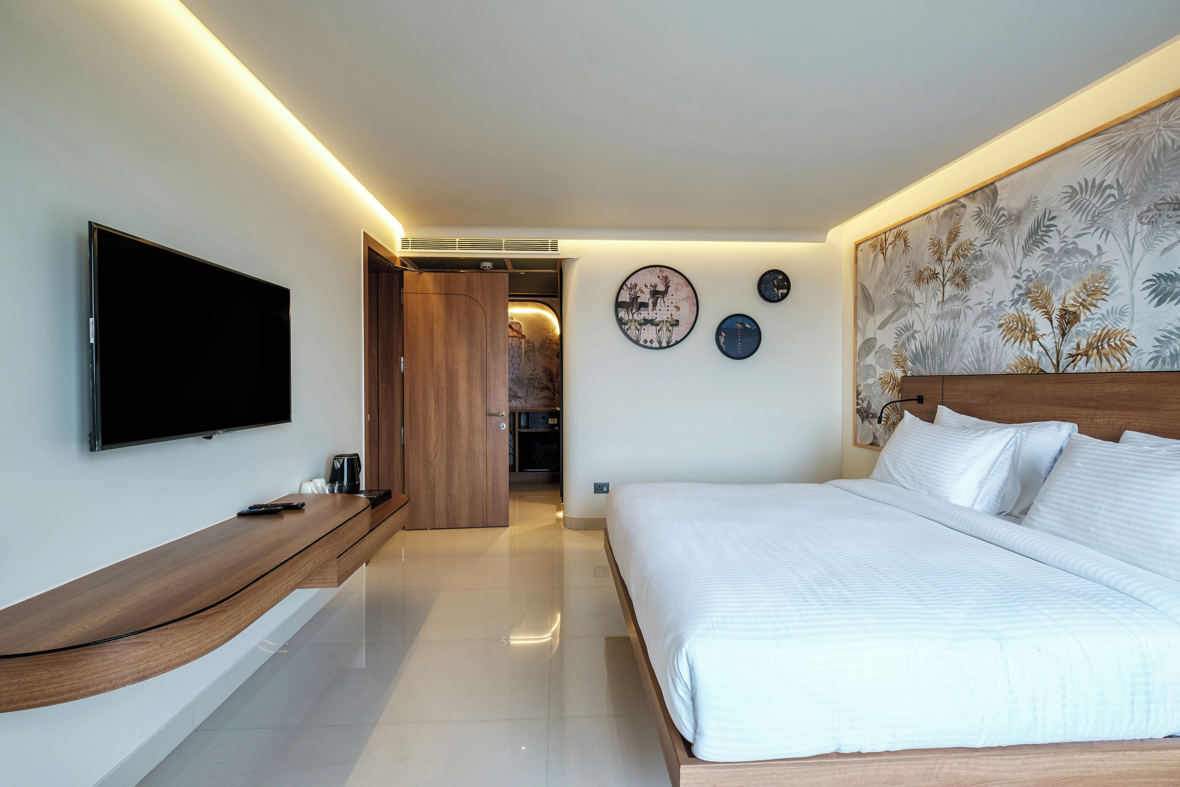A modern bedroom with a large bed, a flat-screen TV, a wooden bench, and a hallway leading to a bathroom.