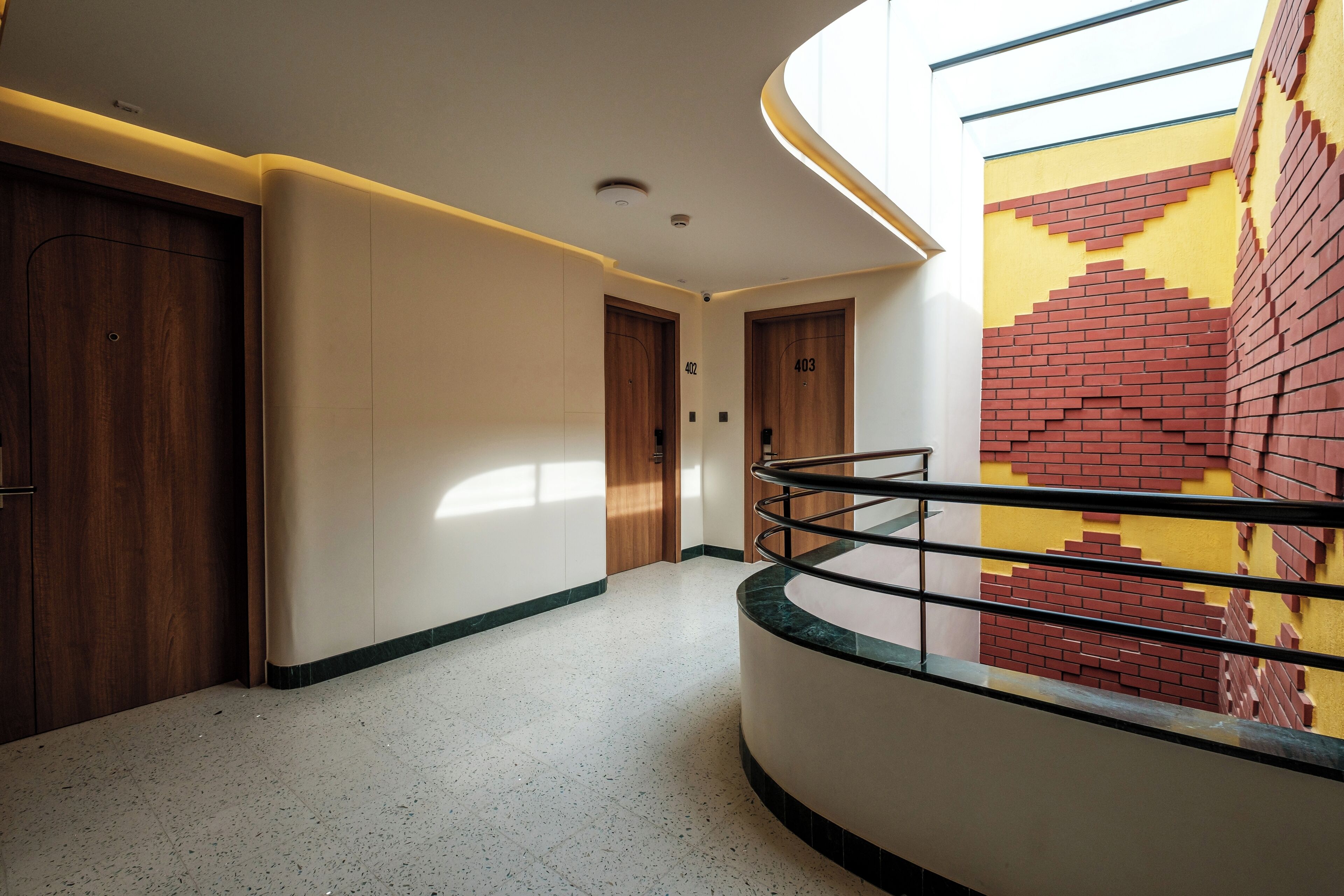 A modern hotel corridor with a curved railing, wooden doors, and a unique brick-patterned wall.