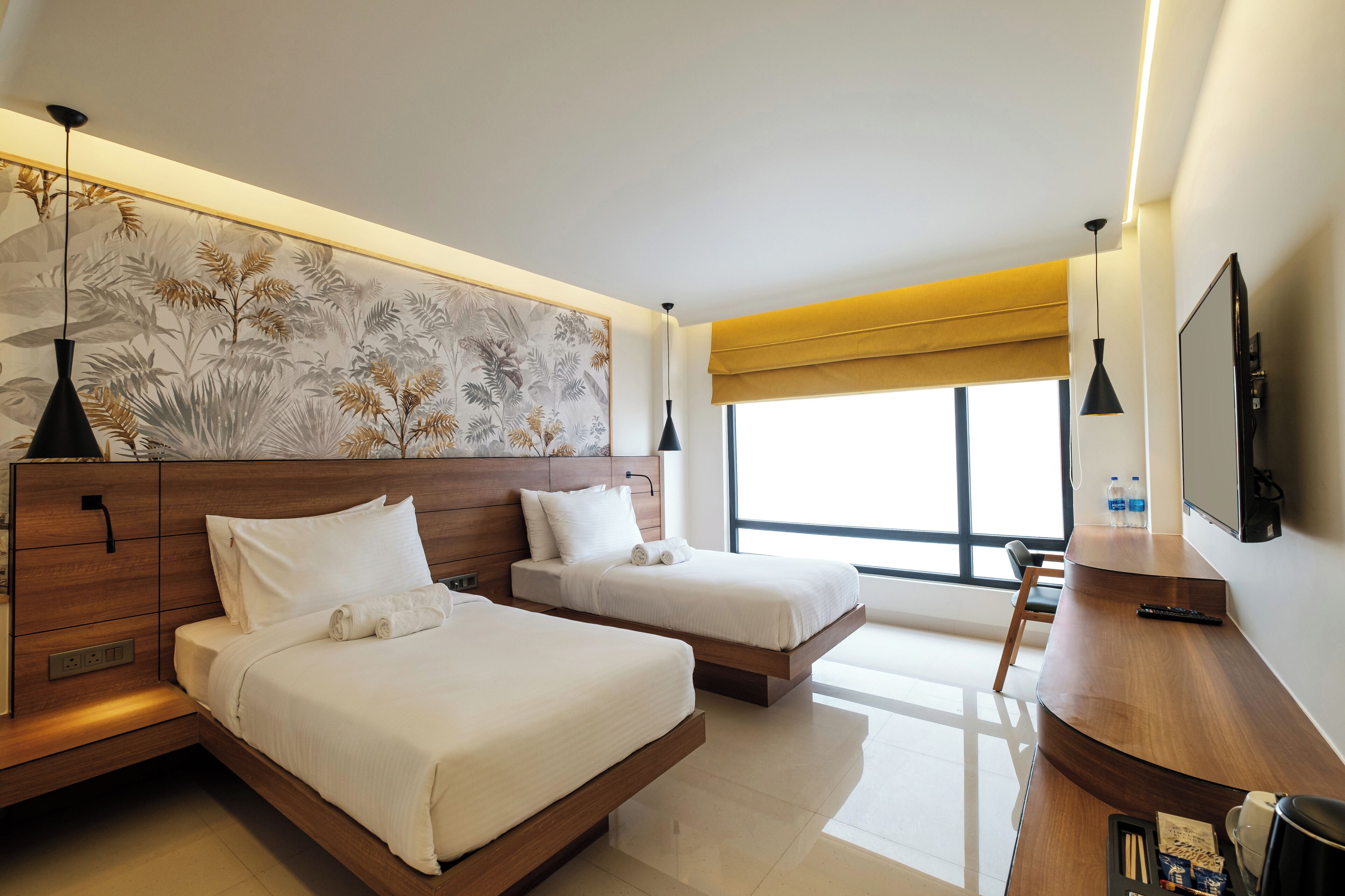 A modern hotel room with two beds, a wooden desk, a large window, and a wall-mounted TV.