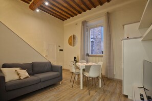 A compact living space with a sofa, dining table, chairs, a TV, and a window with curtains. - Casina Di Giada (Siena)