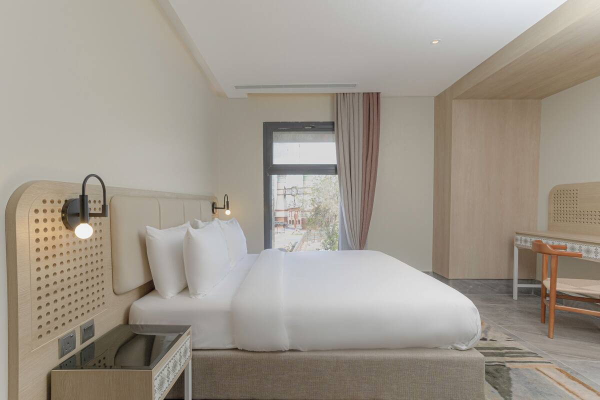 A modern hotel room with a large bed, a wooden headboard, a desk with a chair, and a view of the outside.