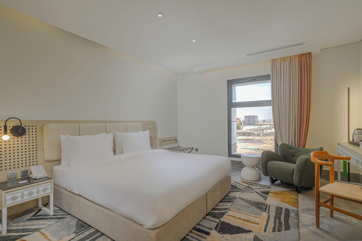 A hotel room with a large bed, a desk with a telephone, a chair, and a view of the city through a window.