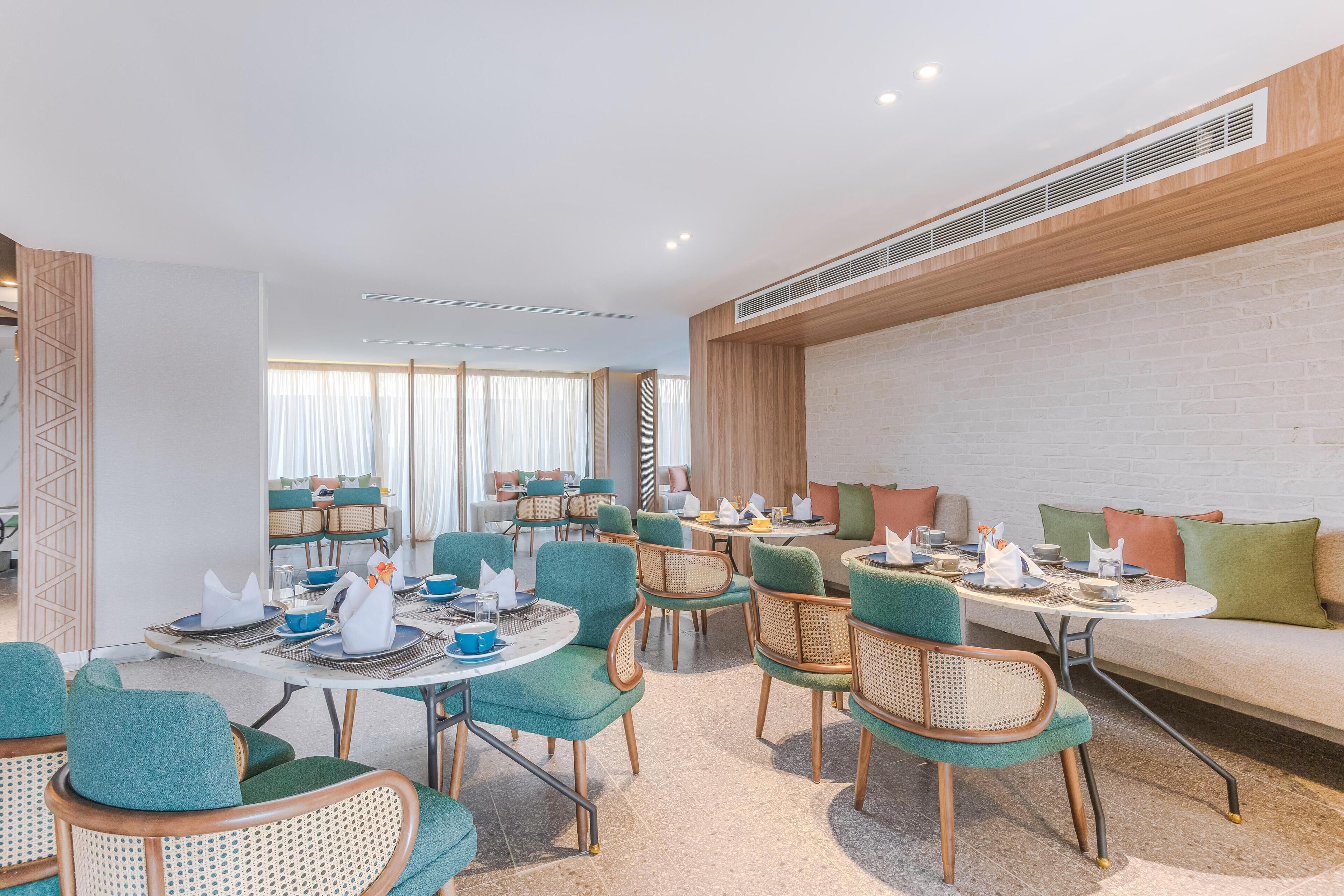 Uma sala de jantar de hotel moderna, com mesas redondas, cadeiras cor turquesa e um sofá com almofadas.