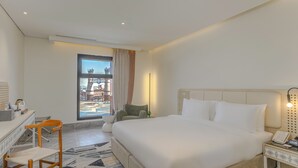 A hotel room with a large bed, a desk with a chair, a nightstand with a phone, and a window with a view of buildings. - Ewaa Express Hotel-Al Shati (Jeddah)