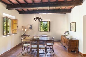 Comedor con una mesa de madera, sillas y una araña. Se ve vegetación por la ventana.