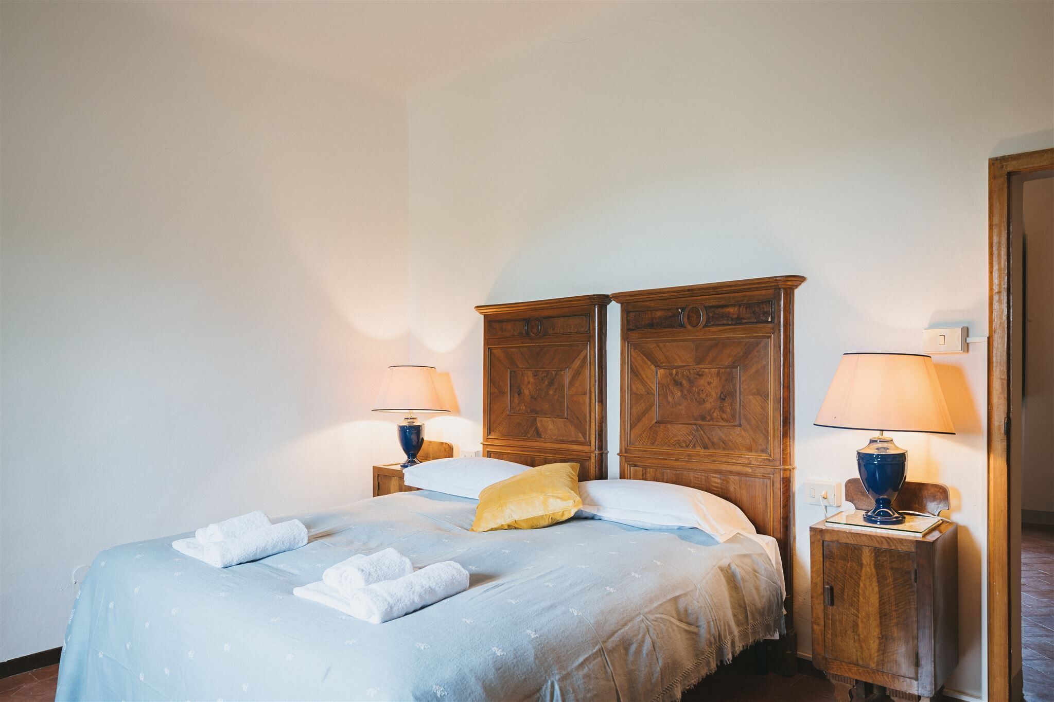 A neatly made bed with a blue bedspread, two pillows, and a set of towels. There are two wooden nightstands with lamps and a wooden wardrobe in the background.