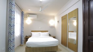 A bedroom with a bed, a ceiling fan, an air conditioning unit, a nightstand, a mirror, and a door. - Canwin Hotel (Accra)