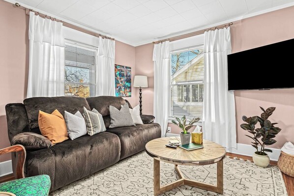 A living room with a dark brown sofa, multiple cushions, a round coffee table, and a flat-screen TV mounted on the wall.