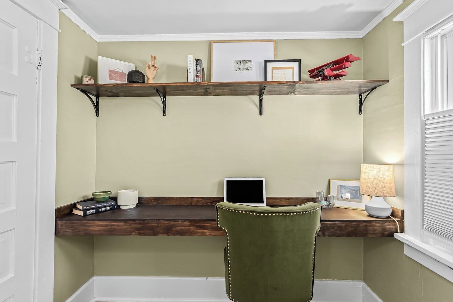 A corner office with a desk, chair, and shelves with books and decorative items.