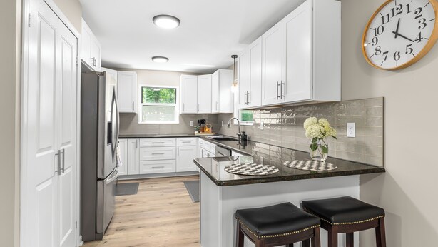 A modern kitchen with white cabinets, a stainless steel refrigerator, a large island with stools, and a wall clock. - Modern [3BR/2BA] Family Home in Commerce Township (Commerce Township)