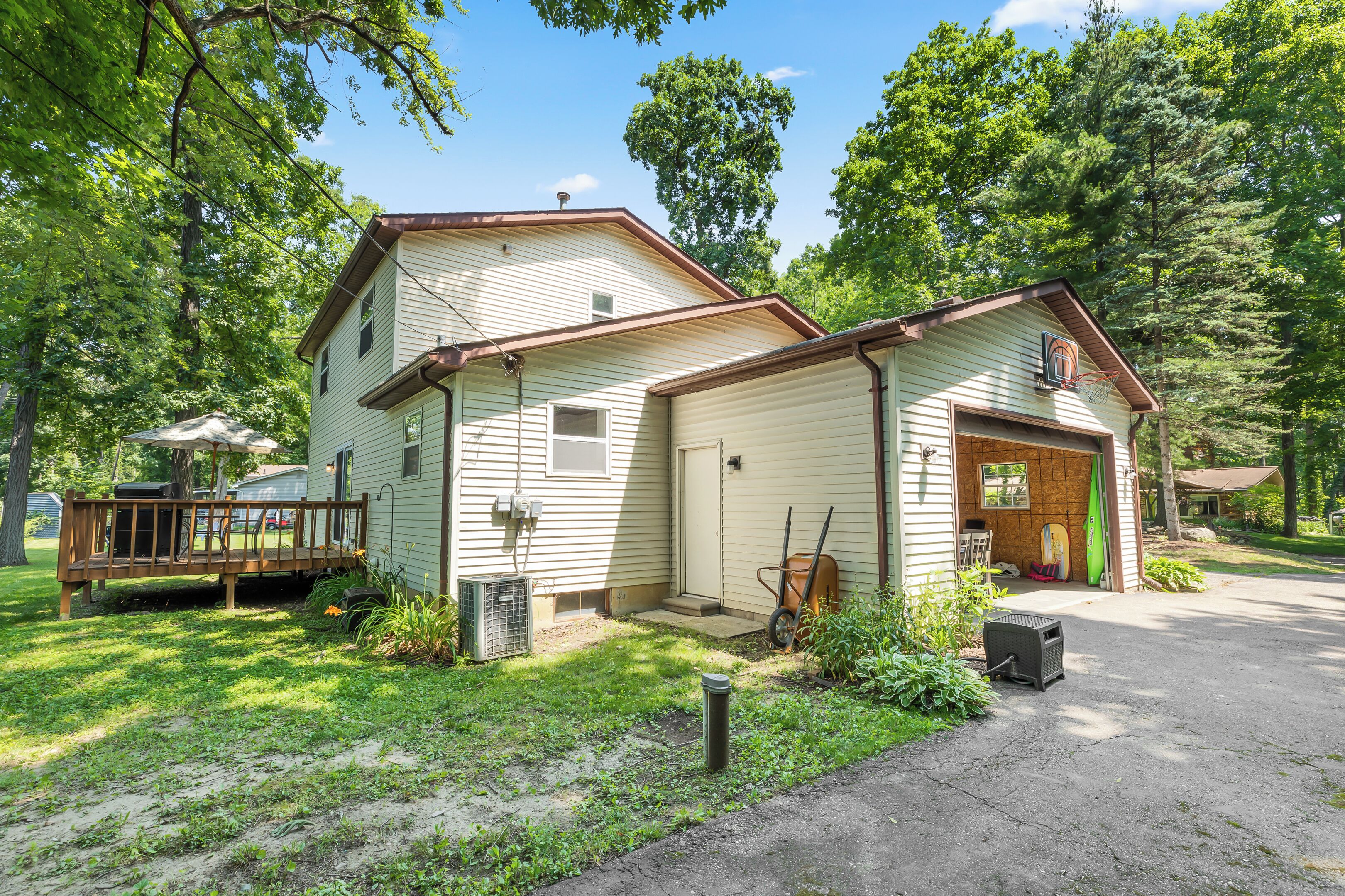 Une maison à deux étages avec un garage, une véranda et un petit jardin où se trouvent une tondeuse à gazon et une brouette.