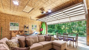 A cozy living room with a sectional sofa, a dining table with chairs, a ping pong table, and a garage door leading to a green outdoor area. - Modern [3BR/2BA] Family Home in Commerce Township (Commerce Township)