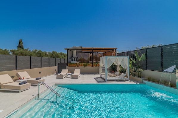 Un espace piscine extérieur moderne, doté de chaises longues, d’un auvent et d’une cascade décorative.