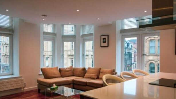 Ein modernes Wohnzimmer mit einer Eckcouch, einem Glastisch und einem Essbereich mit weißem Tisch und Stühlen. Große Fenster sorgen für natürliches Licht und Blick auf die Stadt.