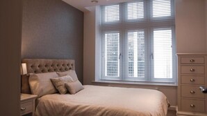 Ein Schlafzimmer mit großem Fenster, einem gepolsterten Kopfteil, einem Nachttisch und einer Kommode.