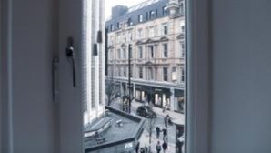 Blick durch ein Fenster eines mehrstöckigen Gebäudes auf eine Straßenszene mit Fußgängern und parkenden Autos.