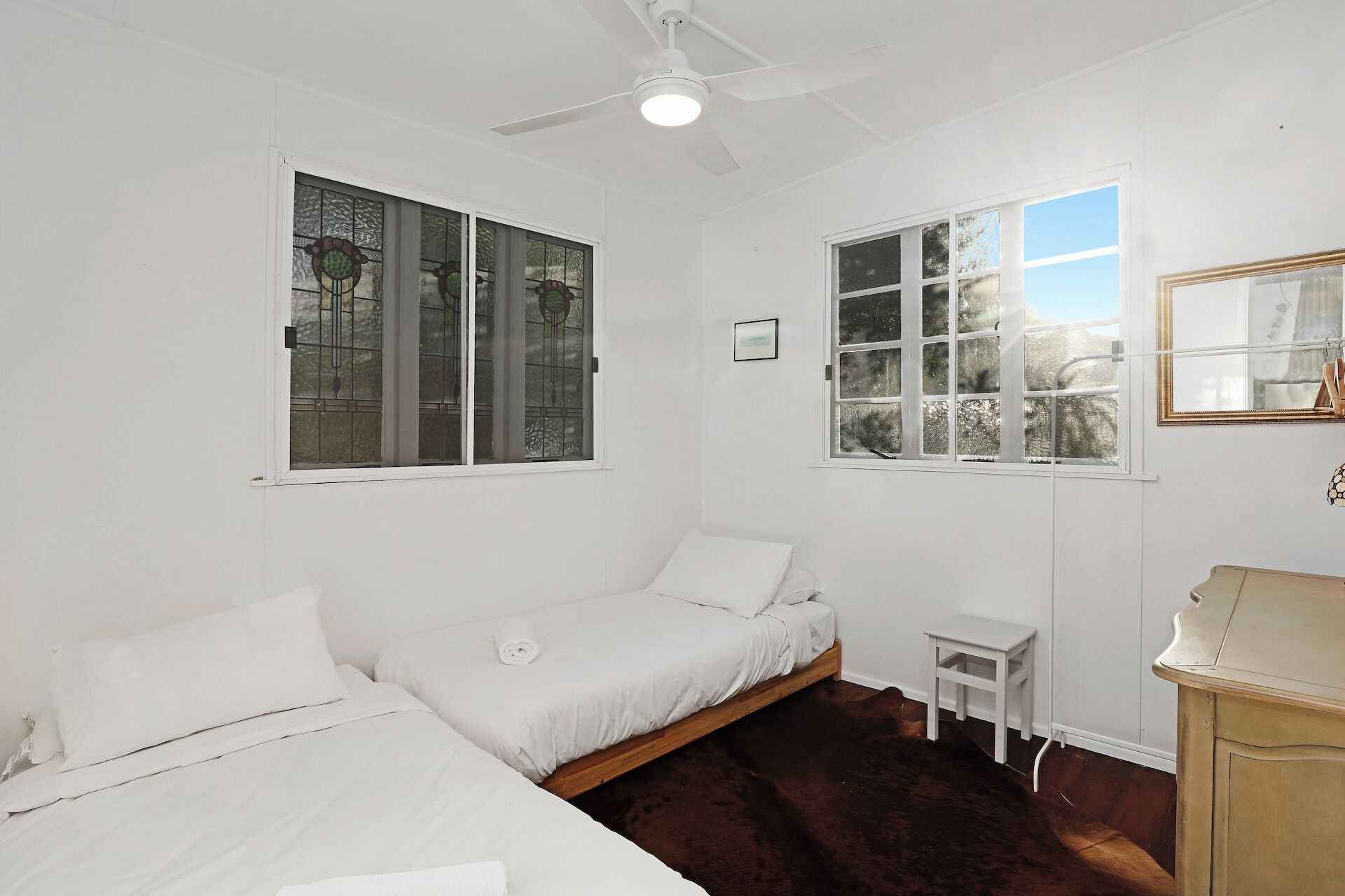 A room with two beds, a small table, a ceiling fan, and a window with a view of trees.