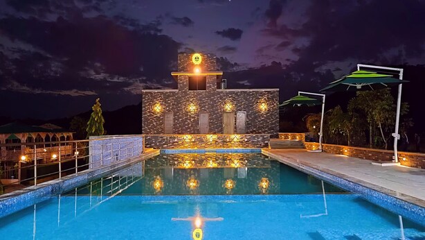 A nighttime view of a swimming pool area with a stone building and a bridge. - The Sky Imperial  Nakoda Green Valley  (Nathdwara)