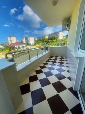 A balcony with a checkered floor, a railing, and a view of buildings and greenery in the background. - Seabird Suite Hotel (Arsin)