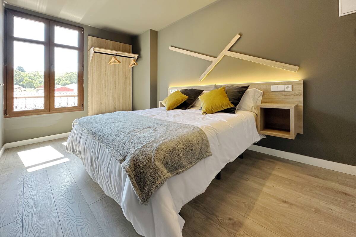 A modern bedroom with a large bed, wooden headboard, and a view of the outdoors through a window.