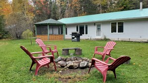 Uma casa com varanda envidraçada, lareira e quatro cadeiras Adirondack no jardim.
