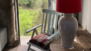 A rustic room with a wooden chair, a small round table, and a red lamp. A window reveals a view of green fields and trees.