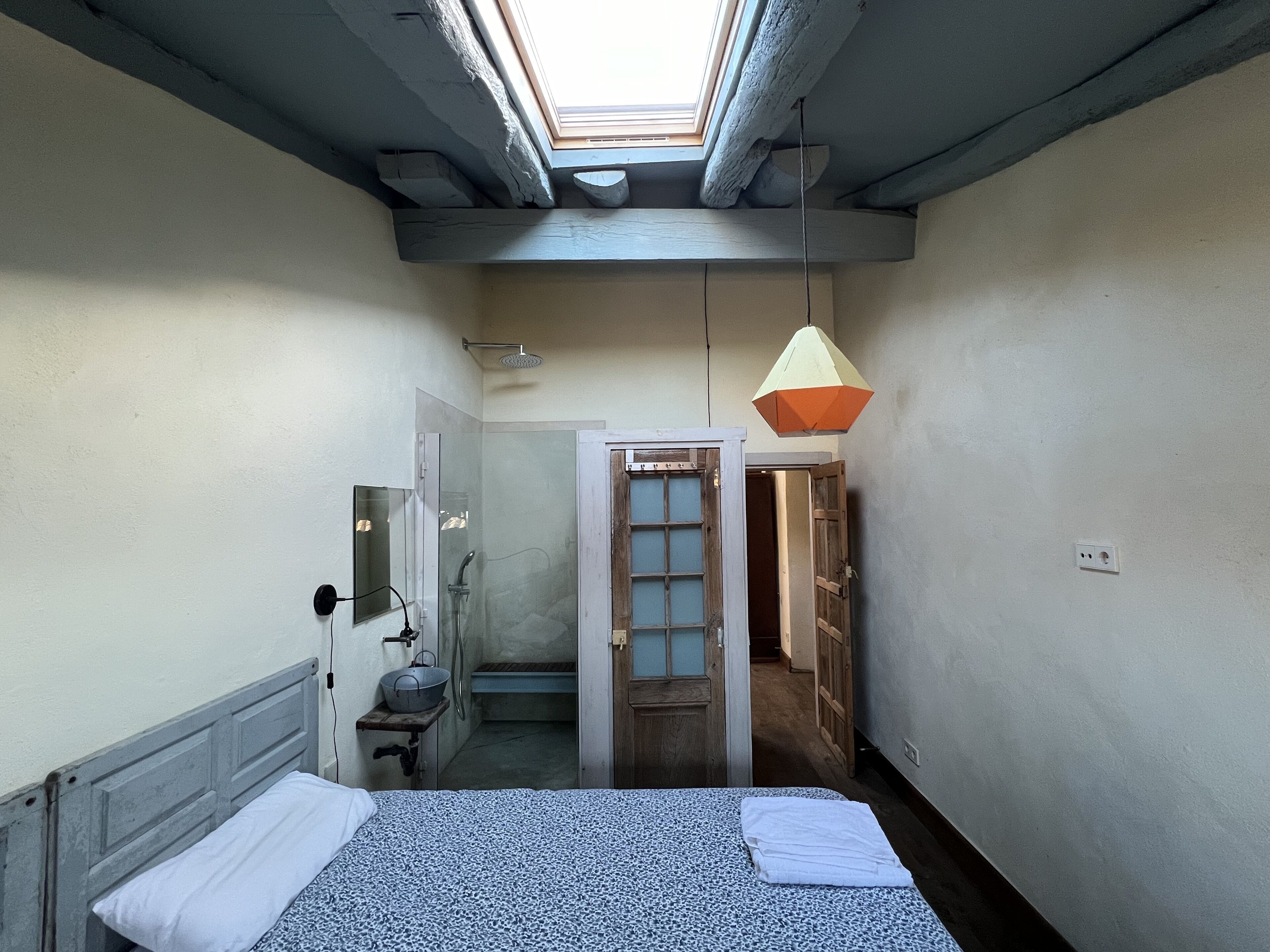 Una habitación estrecha con tragaluz, una cama con colcha estampada, una mesita de noche con lavamanos y una lámpara colgante.