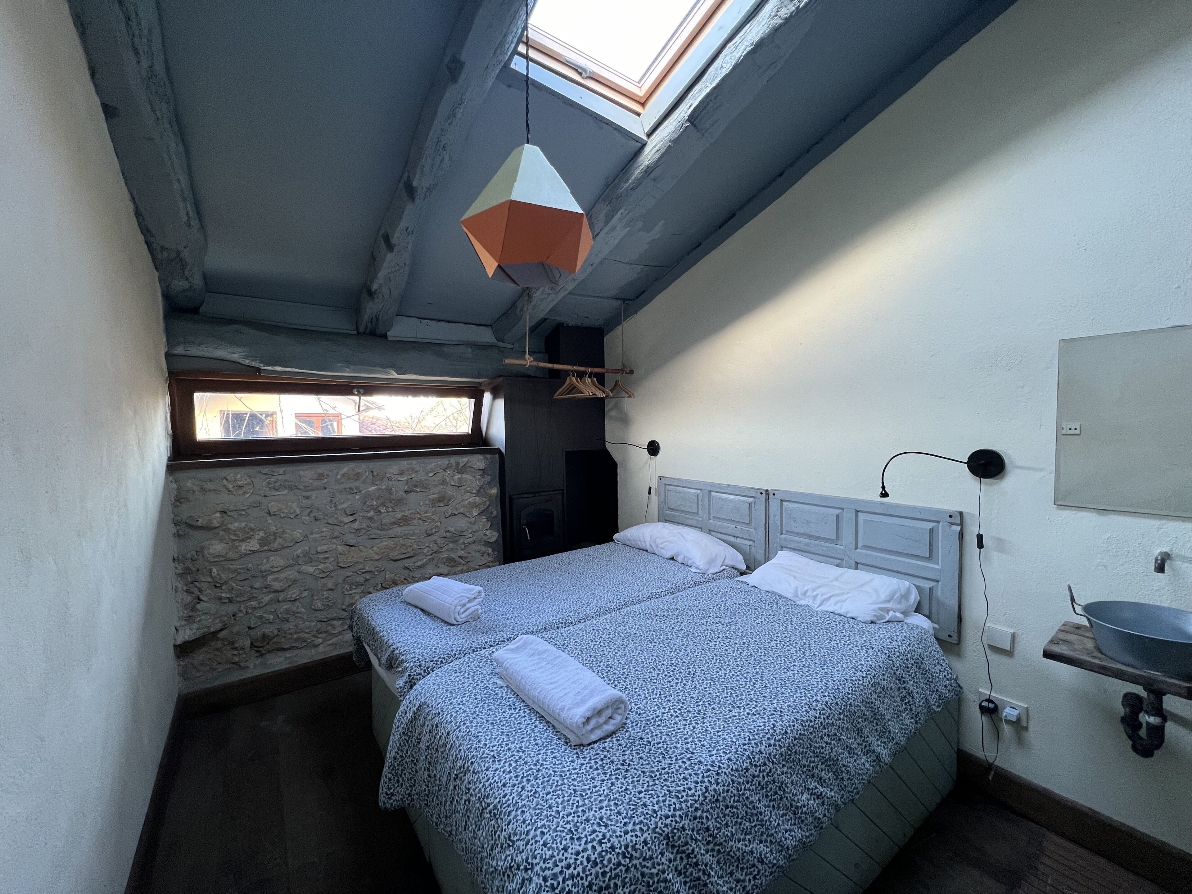 Un dormitorio con dos camas, un tragaluz, un muro de piedra y un lavamanos pequeño.